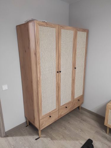 Собран шкаф с тремя дверцами, оснащённый плетёными вставками. Установлены ножки, прикреплены ручки, выровнены элементы. Готовый предмет интерьера стоит на полу.