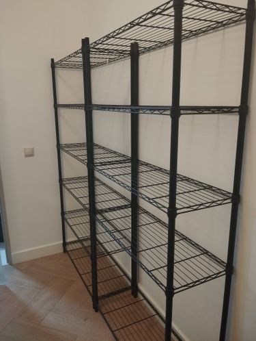 Собрана металлическая конструкция с полками, установлена в углу комнаты. Все элементы надежно закреплены, обеспечивая устойчивость и функциональность для хранения вещей.