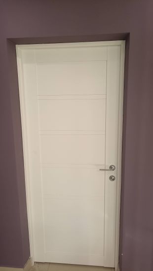 Установлена новая белая дверь, закреплены петли, врезан замок, ручка прикреплена. Проем оформлен наличниками, все элементы подогнаны, конструкция функционирует плавно и надежно.