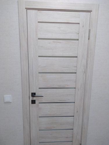 Установлена светлая деревянная дверь с горизонтальными вставками, закреплена на петлях, добавлена ручка и замок, завершена отделка наличниками, обеспечена плавная работа конструкции.