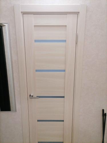 Установлена светлая дверь с горизонтальными вставками, аккуратно закреплена в проеме. Ручка и петли надежно зафиксированы, конструкция ровно выровнена, обеспечивая плавное открытие.
