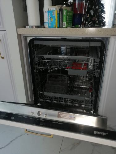 Изображена посудомоечная машина с открытой дверцей на кухне. На столешнице расположены различные предметы, включая коробки и кружку. Машина готова к использованию.