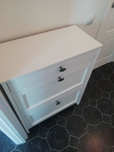 Собран новый белый шкаф с тремя ящиками, установлены ручки, проверена устойчивость и плавность выдвижения. Установлен на черный плиточный пол в светлой комнате.