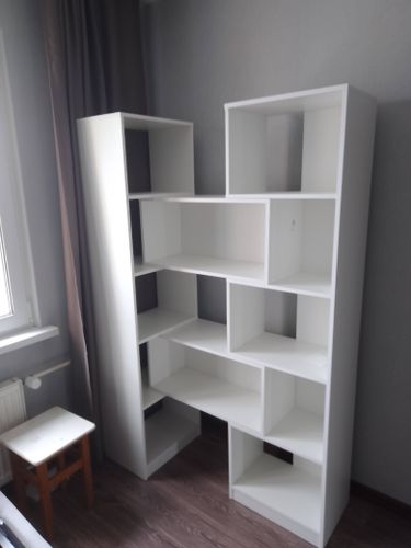 Собран белый стеллаж с асимметричными полками, установлен в комнате у окна. Конструкция устойчива, выполнена аккуратно, все элементы надежно закреплены, готова к использованию.