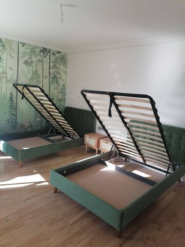 На изображении завершена сборка двух кроватей с подъемными механизмами. Установлены деревянные рамы, обитые зеленой тканью, на полу из светлого дерева. На заднем плане — лесной пейзаж.