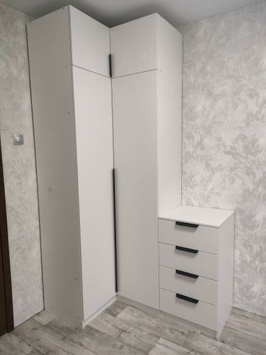 Собран белый комод с четырьмя ящиками, установлен в углу комнаты. Все детали плотно закреплены, ручки прикручены, конструкция устойчива. Пол выполнен в светлых тонах, стена с узором.