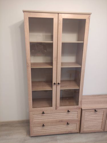 Собран деревянный шкаф с двумя стеклянными дверцами и двумя выдвижными ящиками. Установлены полки, закреплены ручки. Конструкция устойчива и готова к использованию.