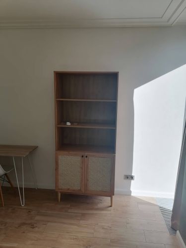 Собран деревянный стеллаж с полками и дверцами, установлен на ножки. Готовое изделие размещено у стены, рядом стол и стул. Пол выполнен из светлого дерева.