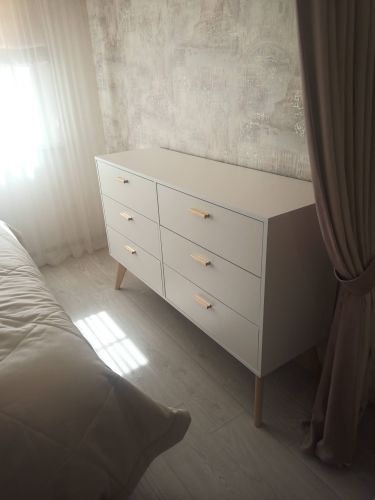 Собран белый комод с шестью ящиками, установлен на деревянные ножки. Все элементы аккуратно подогнаны, ручки прикреплены, конструкция устойчива, размещена у стены в светлой комнате.