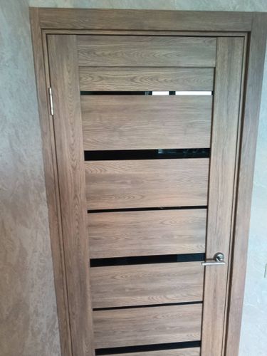 Установлена новая деревянная дверь с вертикальными полосами, закреплены петли, врезана ручка, отрегулированы зазоры, добавлены наличники, обеспечена плавная работа конструкции.