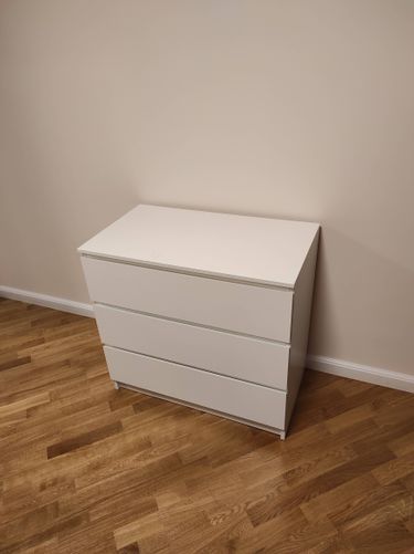 Собран белый комод с тремя ящиками, установлен на деревянный пол у стены. Все элементы надежно закреплены, обеспечивая устойчивость и функциональность мебели.