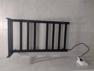 На стене ванной комнаты установлен черный полотенцесушитель с подключением к электросети. Устройство надежно закреплено на плитке, провод аккуратно подключен к розетке.