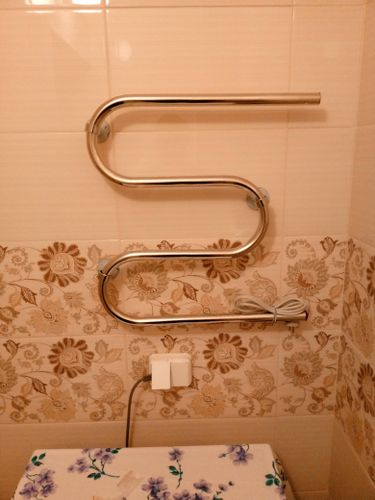 На стене ванной закреплен металлический прибор, подключенный к розетке. Кабель аккуратно уложен, устройство надежно зафиксировано, готово к эксплуатации.