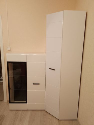 Собран белый шкаф с дверцами, ящиками и стеклянной вставкой, установлен в углу комнаты на светлом полу. Все элементы надежно закреплены, ручки прикручены, конструкция выровнена.