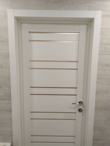 Установлена новая белая дверь с металлической ручкой и горизонтальными вставками. Коробка закреплена, створка ровно навешена, замок функционирует. Завершено оформление проема.