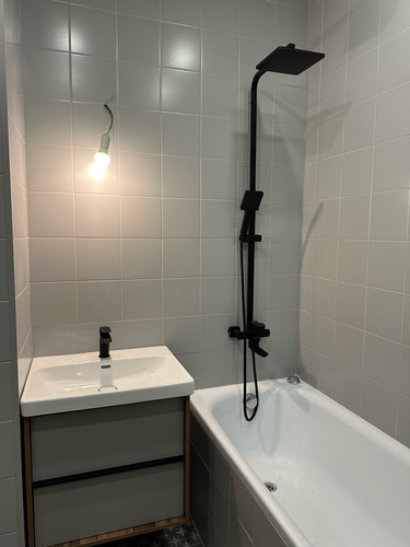 Изображение ванной комнаты с установленным черным душем и смесителем над белой раковиной. Светлая плитка на стенах, горит лампочка. Современный минималистичный стиль.