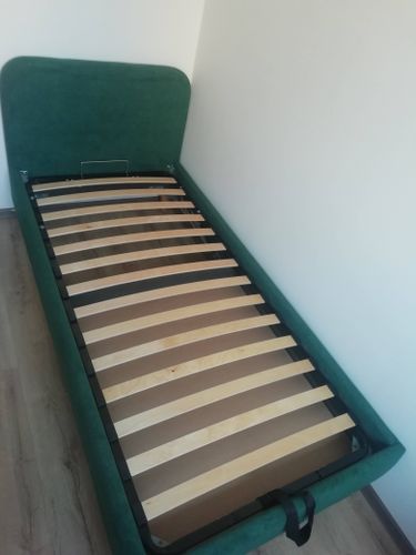 Изготовлена кровать с деревянными ламелями и зелёным изголовьем. Каркас установлен на полу, все элементы надёжно закреплены, конструкция готова к использованию.