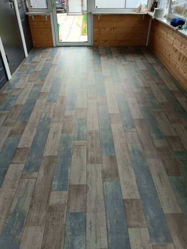 На ровную поверхность уложен линолеум с имитацией деревянных досок, стыки аккуратно подогнаны, покрытие плотно прилегает, создавая эстетичный и практичный пол.