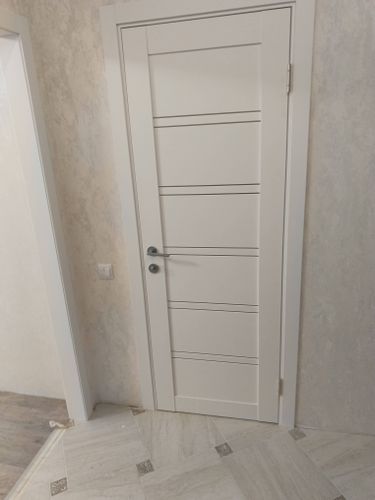 Установлена белая межкомнатная дверь с ручкой и петлями, аккуратно вмонтирована в проем, выровнена по уровню, зазоры минимальны, фурнитура надежно закреплена, отделка завершена.