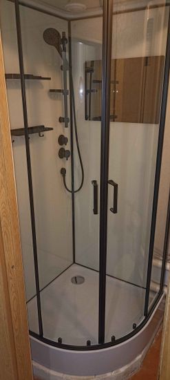 Установлена современная душевая конструкция с прозрачными дверцами и черной рамой. Смонтированы полки, душевой комплект и слив. Завершены все отделочные работы, обеспечена герметичность.