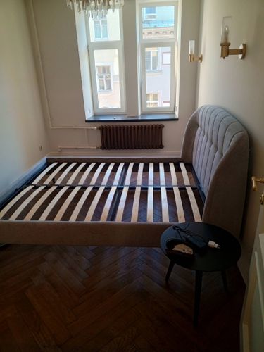 Собран каркас кровати с ламелями, установлено изголовье, прикреплены ножки. Мебель размещена в комнате с паркетом и светлыми стенами. Рядом стоит круглый столик с зарядным устройством.