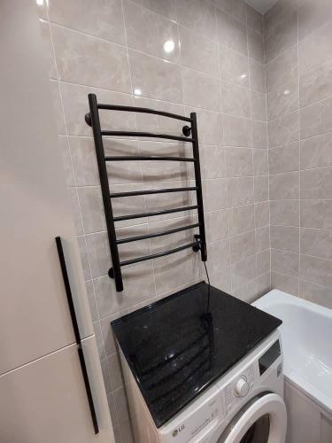 Изображение ванной комнаты с установленной черной лестницей-сушилкой на стене над стиральной машиной с черной столешницей. Стены облицованы светлой плиткой.