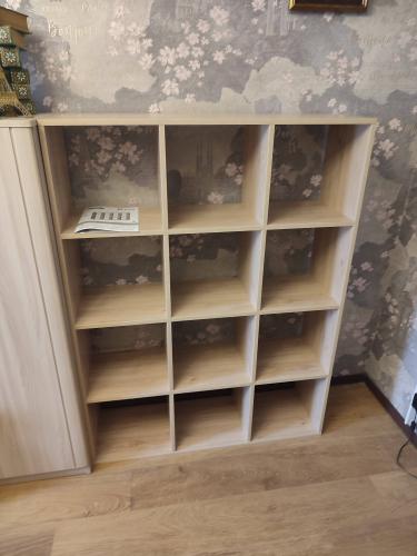 Собран шкаф с девятью отделениями, установлены полки, закреплены детали, проверена устойчивость конструкции. Готовое изделие размещено на полу, рядом с другой мебелью.