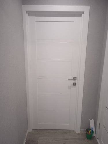 Установлена новая белая дверь с ручкой и замком, выполнена подгонка полотна в проем, закреплены наличники, проведена регулировка петель для плавного закрывания, завершена отделка.