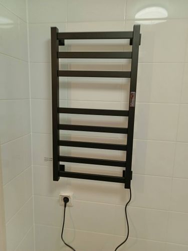 На стене ванной комнаты закреплен электрический прибор для сушки полотенец. Подключен к розетке, провод аккуратно уложен. Устройство готово к использованию, обеспечивая комфорт и тепло.