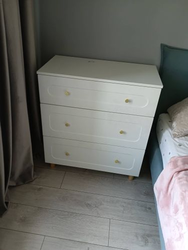 Собран белый комод с тремя ящиками, установлен на ножки, прикреплены ручки, выровнены фасады, проверена устойчивость, размещён у стены рядом с кроватью и шторой.