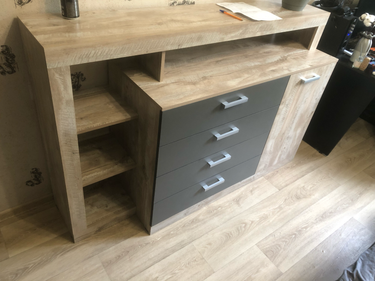 Собран деревянный комод с четырьмя ящиками и двумя полками. Установлены ручки, закреплены детали, проверена устойчивость конструкции. Поверхность выровнена, все элементы на местах.