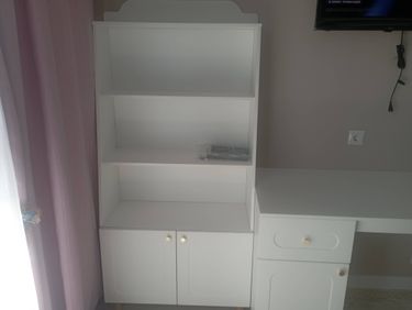 Собран белый стеллаж с полками и дверцами, установлен рядом с письменным столом. Все элементы аккуратно подогнаны, обеспечена устойчивость и функциональность конструкции.