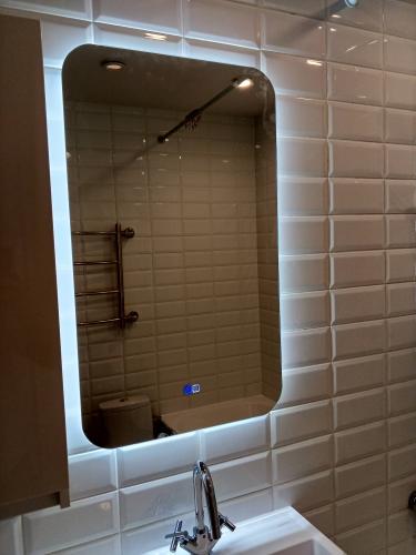 Изображение ванной комнаты с зеркалом, подсвеченным светодиодами, белой плиткой на стенах, раковиной и краном. В отражении видна часть ванной и полотенцесушитель.