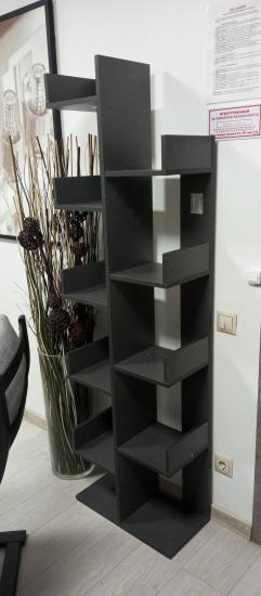 Собран современный стеллаж из темного материала с асимметричными полками. Конструкция устойчивая, установлена в углу комнаты, рядом с декоративными элементами.
