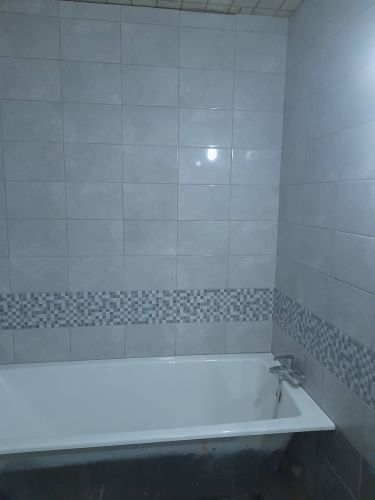 Стены ванной облицованы керамогранитом. Уложен серый кафель с декоративной мозаикой. Поверхность ровная, швы аккуратные, гармонично сочетаются с белой ванной.