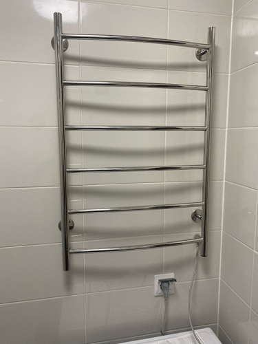 На стене ванной закреплен металлический прибор, подключенный к электросети через розетку. Устройство надежно зафиксировано, провод аккуратно уложен.