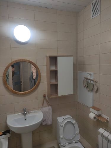 Ванная комната: установлено круглое зеркало с деревянной рамой над раковиной. Закреплено на кафельной стене, обеспечивая устойчивость и безопасность.