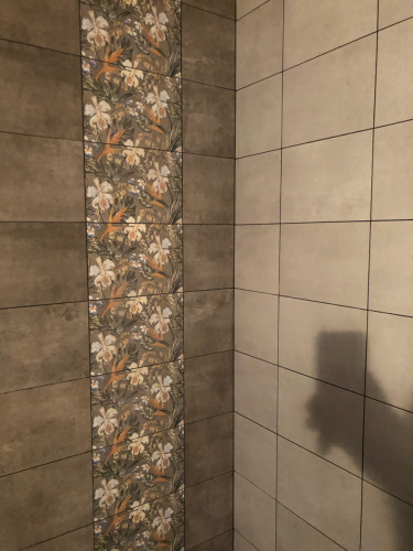 На стене уложен керамогранит с цветочным узором и однотонными плитами. Поверхность ровная, швы аккуратные, создают гармоничный узор. Завершенная работа демонстрирует высокое качество.