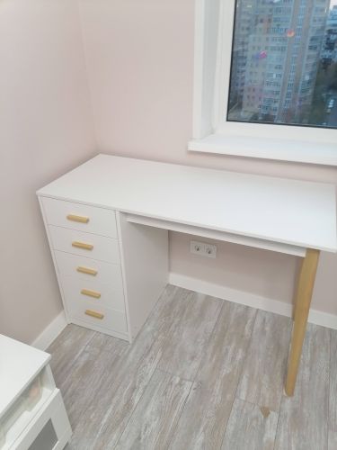 Собран белый комод с четырьмя ящиками и золотистыми ручками. Установлен в углу комнаты на светлом ламинате, рядом с окном. Все элементы надежно закреплены, конструкция устойчива.