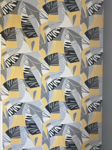 На стене аккуратно наклеены обои с ярким геометрическим узором в серых, желтых и белых тонах. Полотна тщательно совмещены, создавая цельный и гармоничный вид.