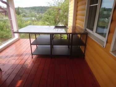 На террасе установлена металлическая конструкция с полками и столешницей. Стол расположен рядом с окном, на фоне зелёного пейзажа. Пол выполнен из деревянных досок.