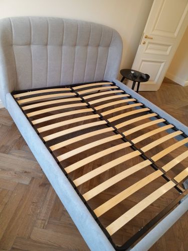 Изображение демонстрирует завершённую сборку кровати с деревянными ламелями на каркасе, установленной на паркетном полу в светлой комнате с открытой дверью и небольшим столиком рядом.