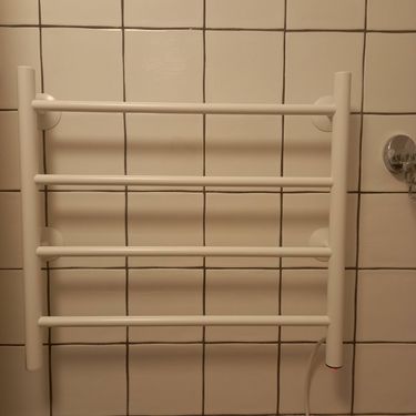 На стене ванной комнаты закреплён электрический прибор для сушки полотенец, подключённый к сети. Устройство установлено ровно, провода аккуратно спрятаны, обеспечивая безопасное использование.