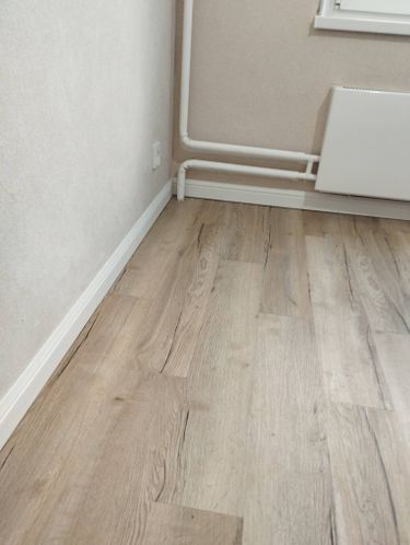 Установлен белый плинтус вдоль стены, аккуратно прилегающий к ламинату. Все стыки ровные, без зазоров. Плинтус гармонично сочетается с интерьером, создавая завершенный вид помещения.