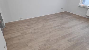 Завершена укладка деревянного покрытия: ровные доски уложены на пол, создавая гладкую поверхность. Светлый оттенок материала гармонично сочетается с белыми стенами, добавляя уют в помещение.