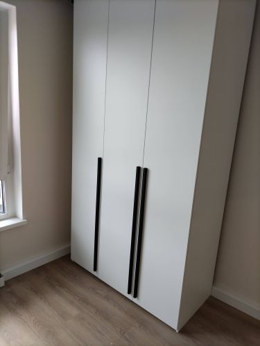 Собран белый шкаф с тремя дверцами и черными ручками. Установлен на деревянный пол рядом с окном. Конструкция завершена, все элементы надежно закреплены.