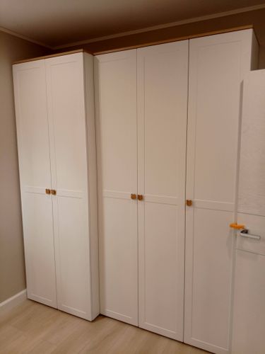 Собран белый шкаф с четырьмя дверцами, аккуратно установлен в углу комнаты. Двери оснащены золотистыми ручками. Конструкция устойчива и гармонично вписывается в интерьер.