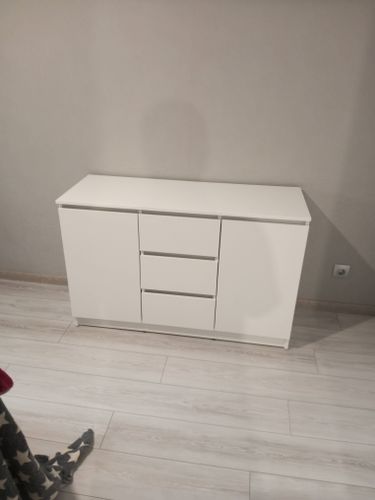 Собран белый комод с тремя ящиками и двумя дверцами, установлен на светлом полу у стены. Все детали соединены, конструкция устойчива, готова к использованию.