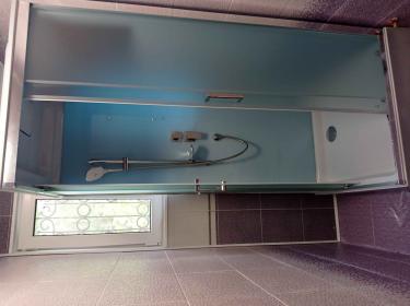 Изображена современная душевая кабина в ванной комнате с фиолетовой плиткой. Установлены стеклянные двери, душевой шланг и смеситель. Окно с решеткой обеспечивает естественное освещение.