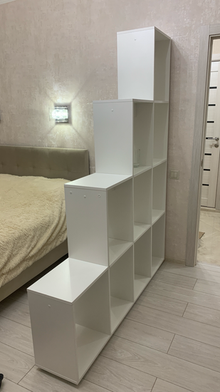 Собран белый стеллаж с девятью ячейками, установлен в спальне. Конструкция устойчива, элементы надежно закреплены, обеспечивая функциональность и эстетичный вид.